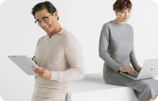 a person sitting on a countertop working on a laptop next to a person, leaning against the same counter, working on a tablet